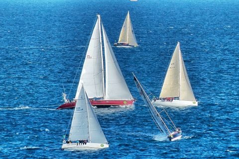 Six racing yachts under white sails on a deep blue sea off Swanage at the start of the 2013 Fastnet Race, one heeled hard over as it tacks