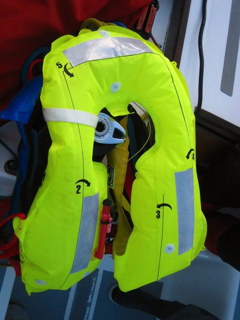 A fully inflated yellow gas lifejacket with reflective patches and a harness ring, hanging on board a boat