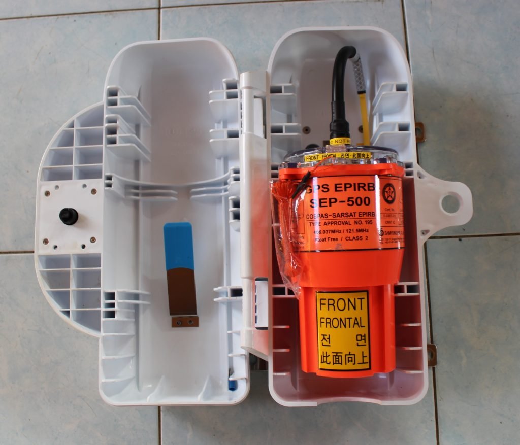 An orange 406 MHz GPS EPIRB sitting in its opened white float-free bracket housing on a tiled floor