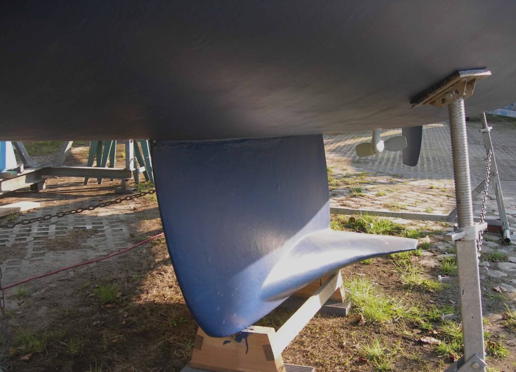 The blue-painted winged keel of a yacht hauled out ashore, resting on a wooden block beside a steel hull support stand