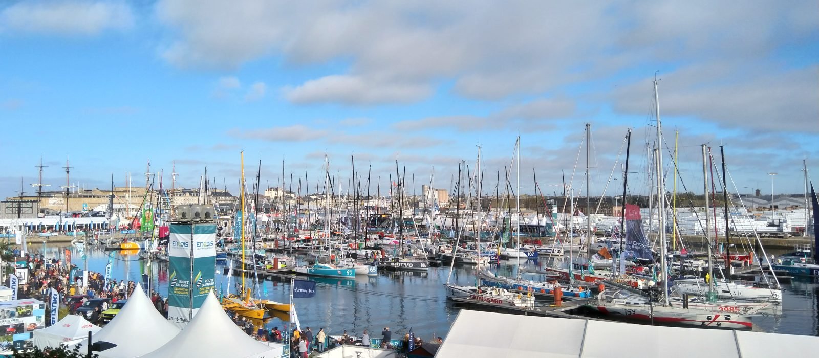Racing multihulls and monohulls moored side by side in the Vauban basin at Saint-Malo with the town behind, crowds walking the quays before a Route du Rhum start