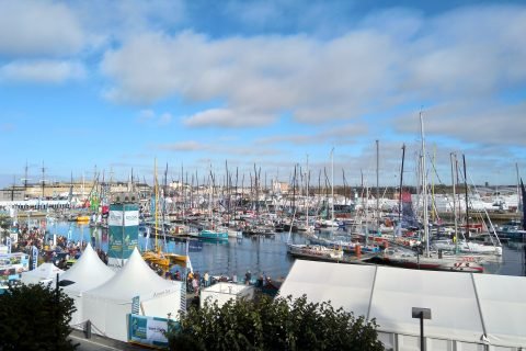 Racing multihulls and monohulls moored side by side in the Vauban basin at Saint-Malo with the town behind, crowds walking the quays before a Route du Rhum start