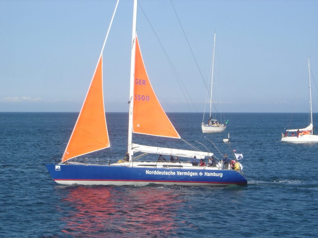 A blue racing yacht sailing under a small orange storm jib and orange storm trysail, mainsail furled on the boom, with two yachts behind